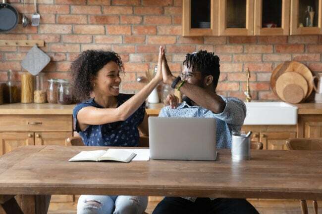 Heyecanlı genç Afrikalı Amerikalı çift ev mutfağındaki masada oturup dizüstü bilgisayarda beşlik çakarak çevrimiçi piyango zaferini kutluyorlar. Mutlu çift ırklı erkek ve kadın bilgisayarda çalışarak başarının tadını çıkarıyor. Şans kavramı.