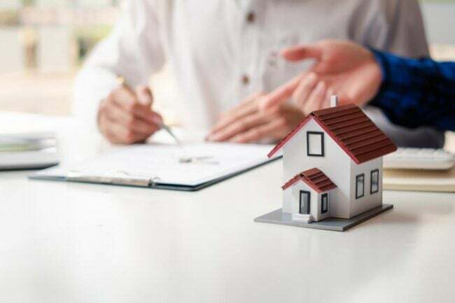 Uma pequena casa modelo branca com telhado vermelho fica sobre uma mesa onde duas pessoas apontam e escrevem em um documento. 