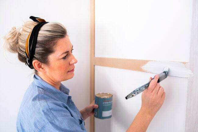Mavi yakalı gömlekli bir kadın küçük bir boya fırçasıyla duvardaki ahşap panellere astar uyguluyor.