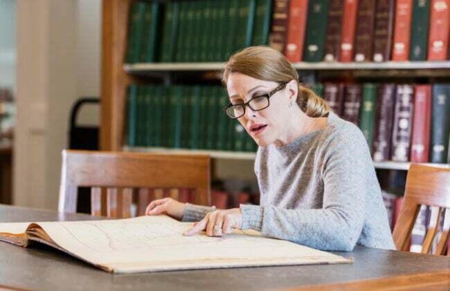 Een volwassen vrouw van in de veertig die onderzoek doet in de bibliotheek, zittend aan een tafel en naar een groot, oud boek kijkt.