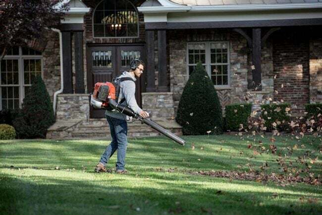 Людина використовує рюкзак для повітродувки листя Husqvarna, щоб перебирати листя у дворі з великим будинком на задньому плані.