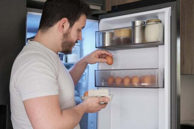 Молодой латиноамериканец кладет яйца на полку двери холодильника.