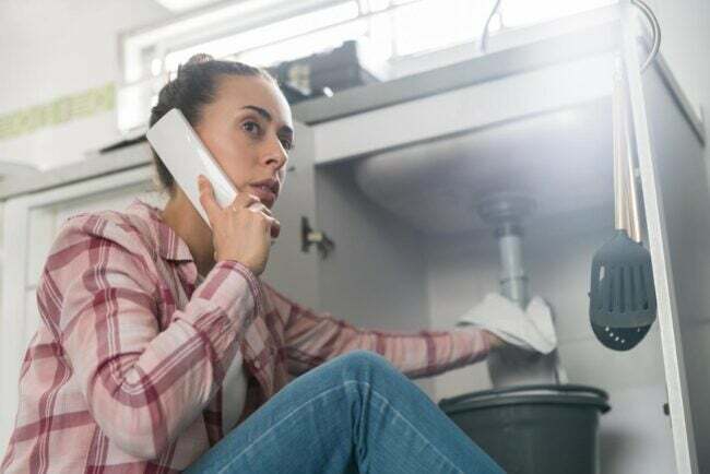 Egy nő telefonál, miközben megpróbálja megállítani a mosogató alatti szivárgást.