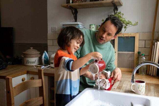 家で一緒に食器を洗ったり乾かしたりしている男児と父親の上半身ショット。 二人とも笑顔でカジュアルな服装をしています。