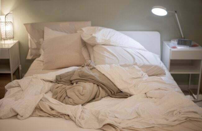 Bej ve beyaz çarşaflı dağınık yatak, yatağın etrafındaki komodinlerde lambalar