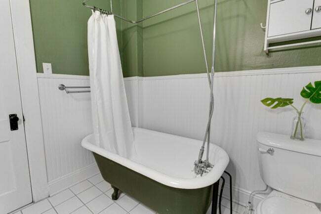 Egyszerű fürdőszoba zöld falakkal, fehér karmos lábfürdővel, zuhanyfüggönnyel és lambériával.