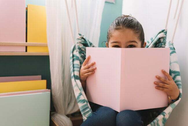 menina escondendo o rosto atrás de um livro rosa em um pequeno canto de leitura