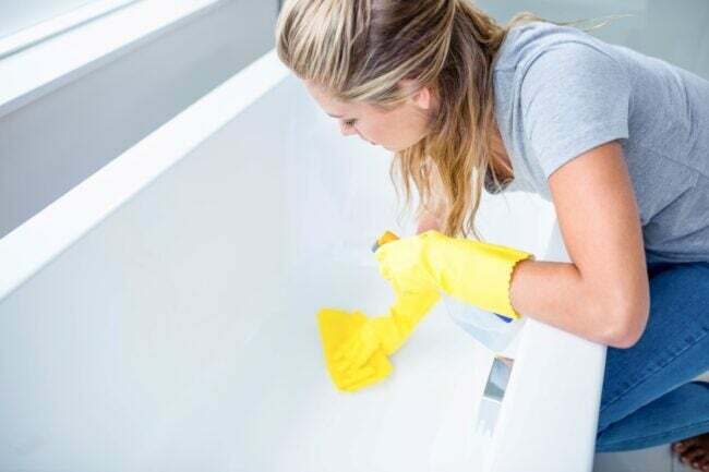 Mulher usando luvas de borracha amarelas limpando o fundo de uma banheira.