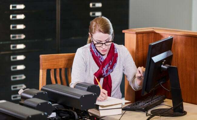 Zrelá žena v knižnici používa počítač na prezeranie historických dokumentov uložených na mikrofiši. Píše si poznámky.