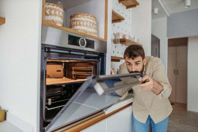barzdotas-vyras-uždegimo marškinėliai-laiko atsuktuvą, o jis žiūri į sieninę viryklę baltoje virtuvėje