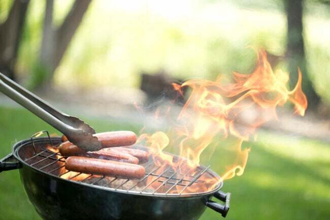 grătar rotund afară, pe gazon verde, cu clești, punând hot-dog-uri pe flacără deschisă