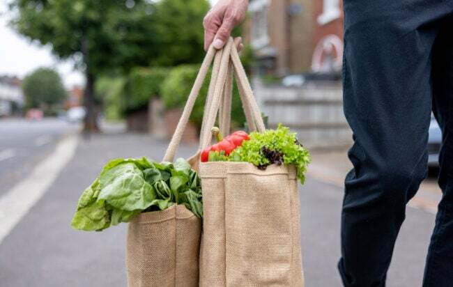 Vyras, gatvėje nešantis daugkartinį pirkinių maišelį su šviežiomis daržovėmis.