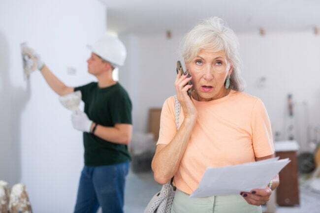 starší žena v oranžové košili mluví do mobilního telefonu, zatímco dodavatel pracuje na bílé zdi za ní 