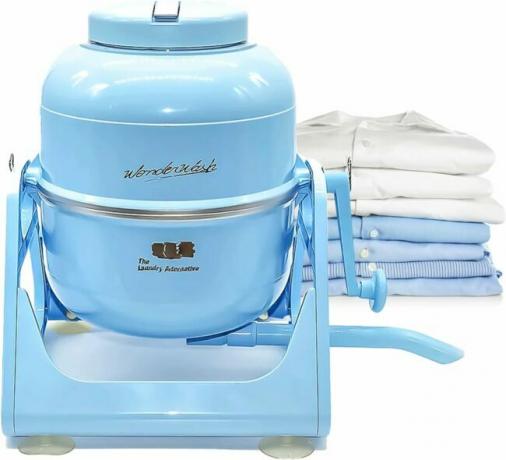 Gaiši zila manuāla veļas mašīna ar rokas kloķi atrodas blakus salocītas veļas kaudzei.