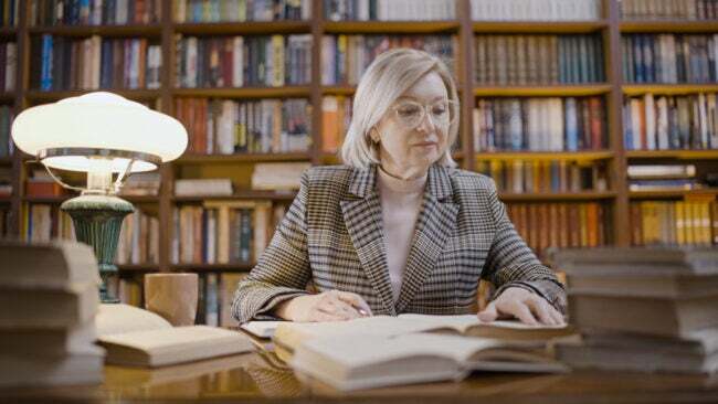 Žena v knižnici si číta knihy na stole pred sebou.