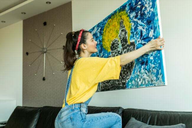 iStock-1311835719 decora cu meșteșuguri femeie care atârnă pictură abstractă albastră și galbenă pe perete