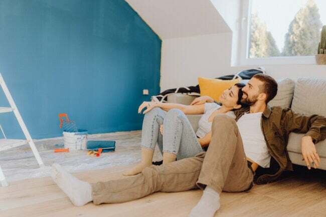 Молодая пара сидит на полу и любуется свежевыкрашенной бирюзовой стеной, которую они покрасили вместе. 