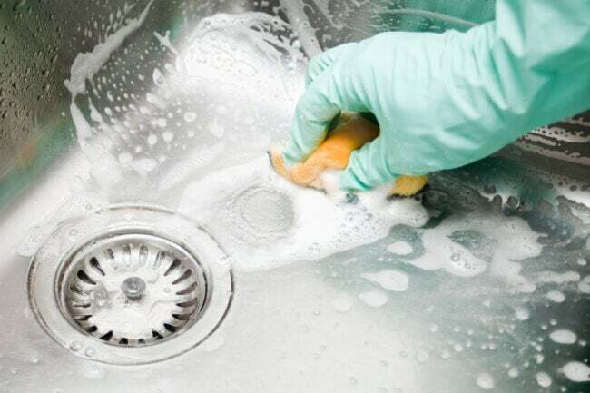 Manos humanas con guantes protectores fregando un fregadero de cocina grande de forma cuadrada con un estropajo, burbujas de jabón.
