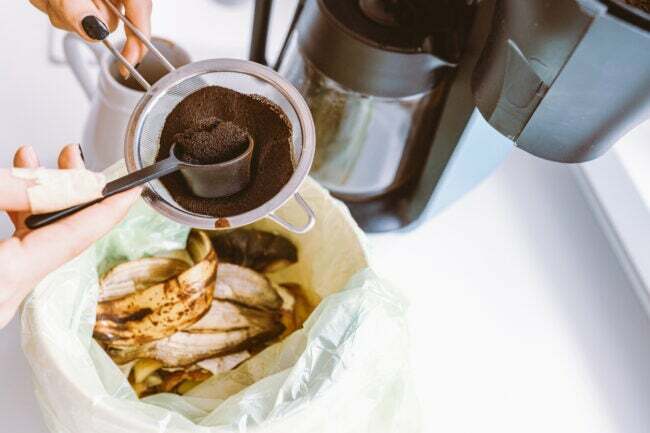 Borras de café usadas sendo colocadas em um recipiente de compostagem ao lado de uma cafeteira. 