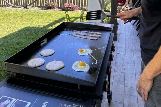 Persona che cucina pancake, uova e pancetta su una piastra Blackstone da 36 pollici nel cortile.